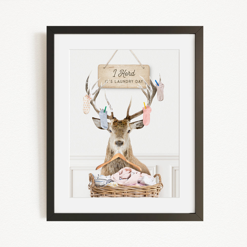 A framed image depicting a deer with antlers wearing a clothesline, with a sign above that reads "I herd it's laundry day".