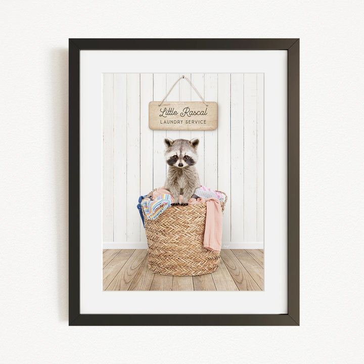 A raccoon sitting in a woven laundry basket, with a sign above that reads "Little Racquel Laundry Service".