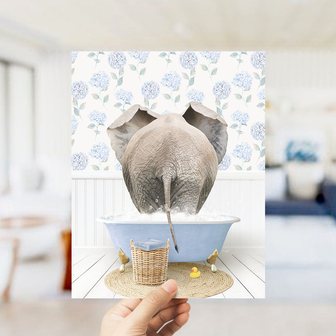 A hand holds up a framed image depicting an elephant standing in a blue bathtub filled with water, surrounded by a floral pattern.