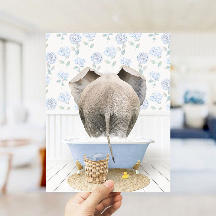 A hand holds up a framed image depicting an elephant standing in a blue bathtub filled with water, surrounded by a floral pattern.