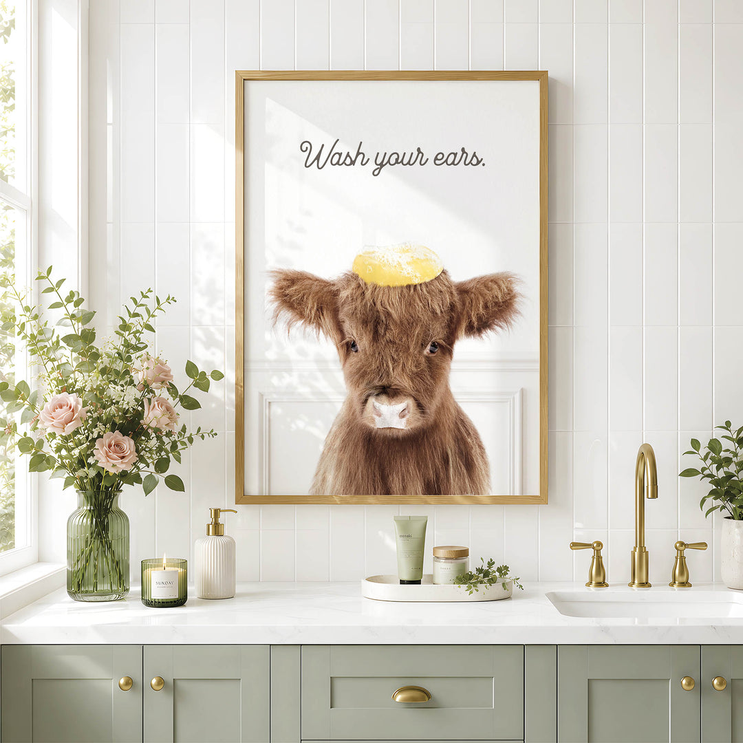 a bathroom counter with a framed picture of a cow wearing a yellow hat, a vase of flowers, and various toiletries such as a soap dispenser and a candle.