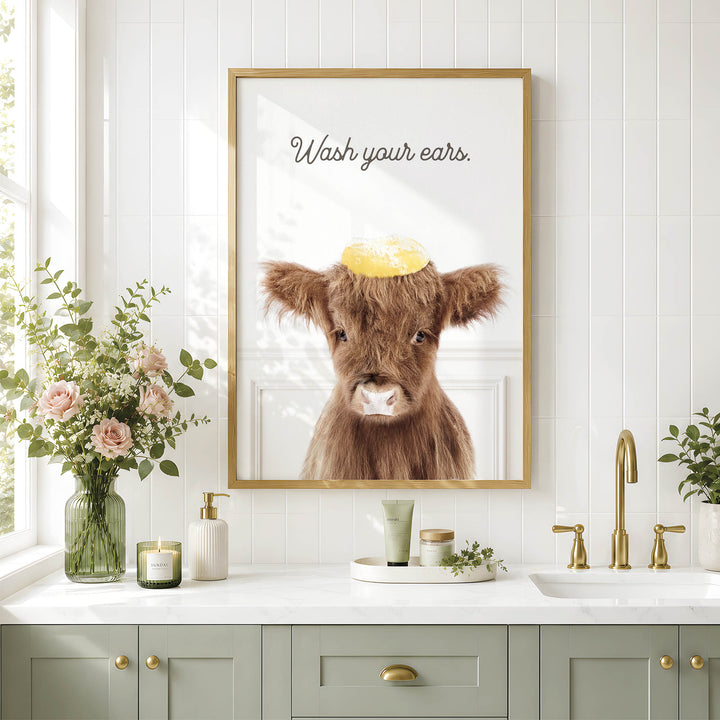 a bathroom counter with a framed picture of a cow wearing a yellow hat, a vase of flowers, and various toiletries such as a soap dispenser and a candle.