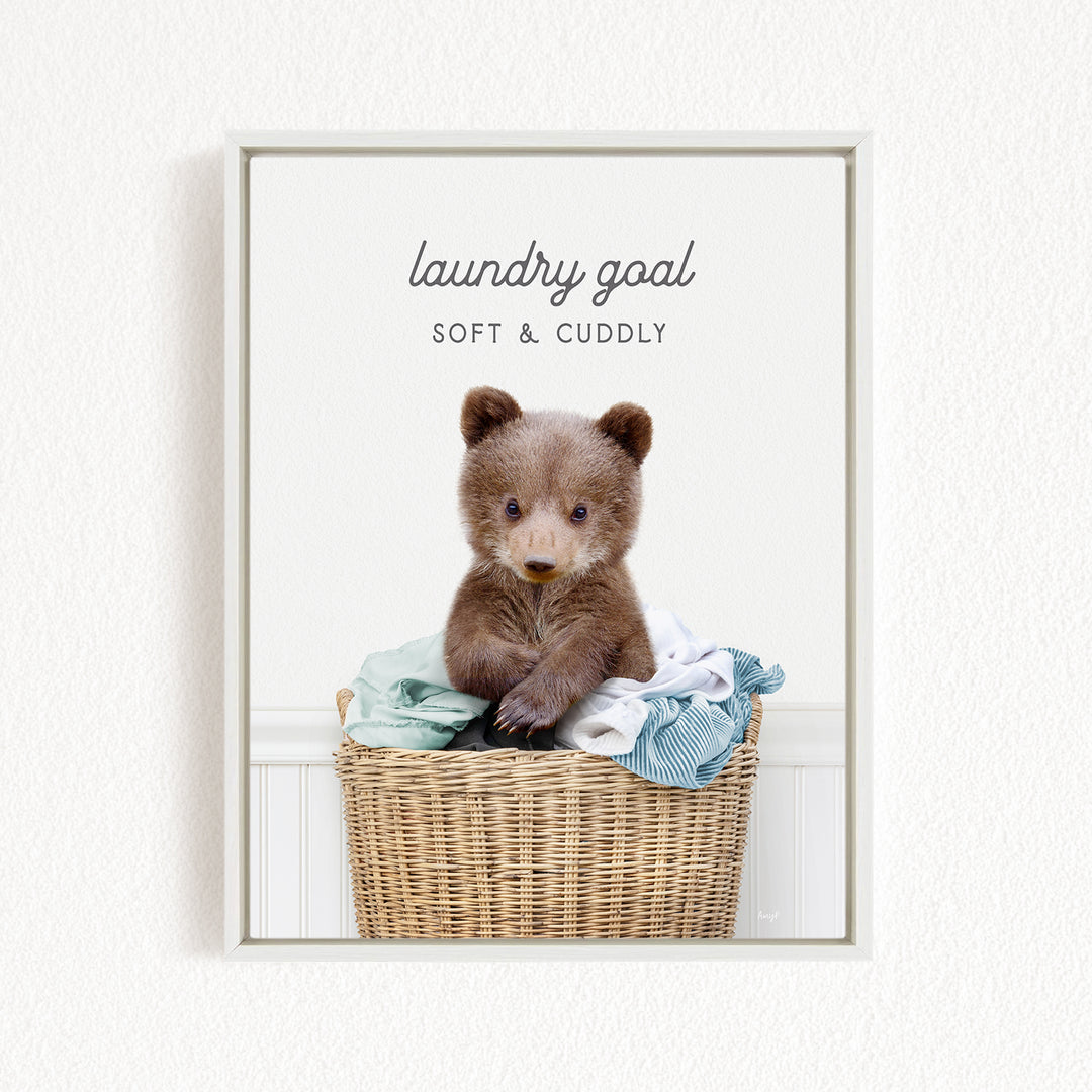 A cute brown bear sitting in a wicker laundry basket filled with clothes, with the text "laundry goal" and "soft & cuddly" written above it.