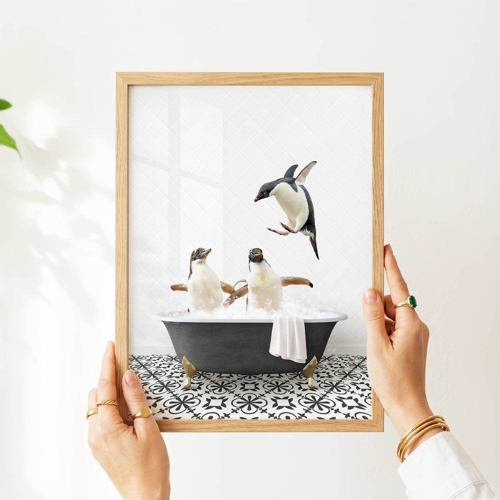 A person's hands are holding a framed picture of two penguins sitting in a bathtub, with one penguin flying out of the tub.