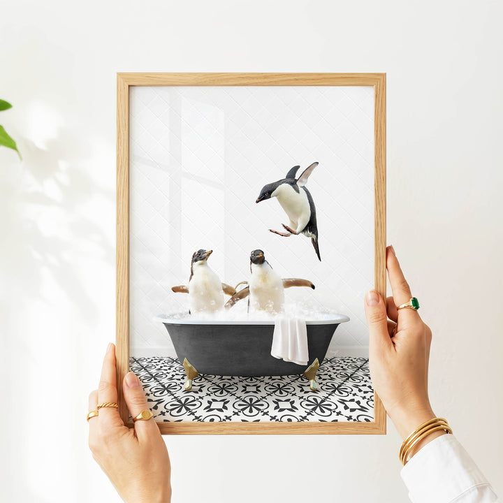 A person's hands are holding a framed picture of two penguins sitting in a bathtub, with one penguin flying out of the tub.
