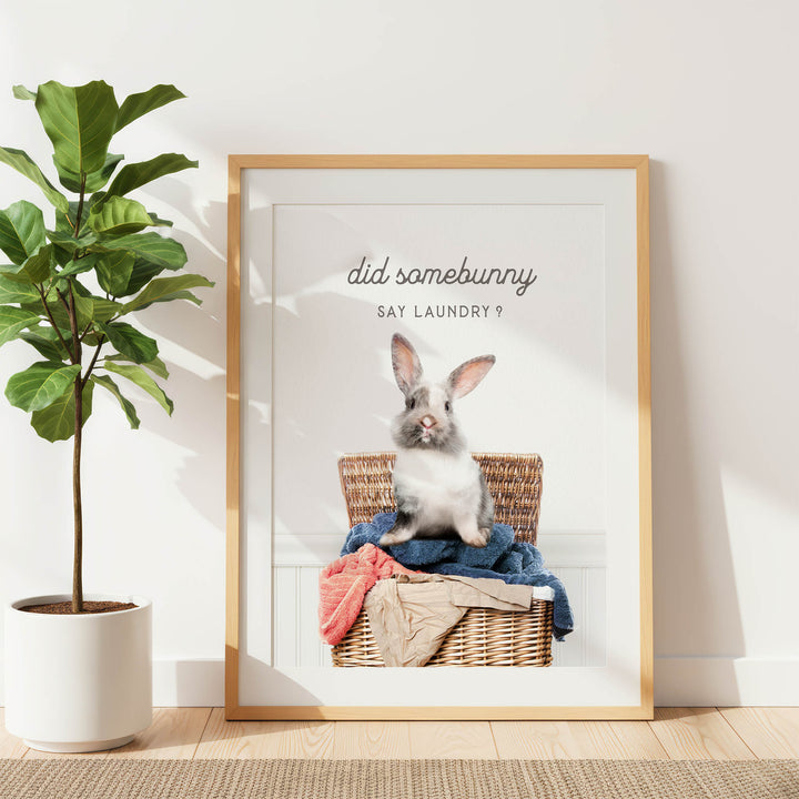 A framed poster with a rabbit sitting in a wicker basket, surrounded by clothes, with the text "did some bunny say laundry?" displayed above the image.