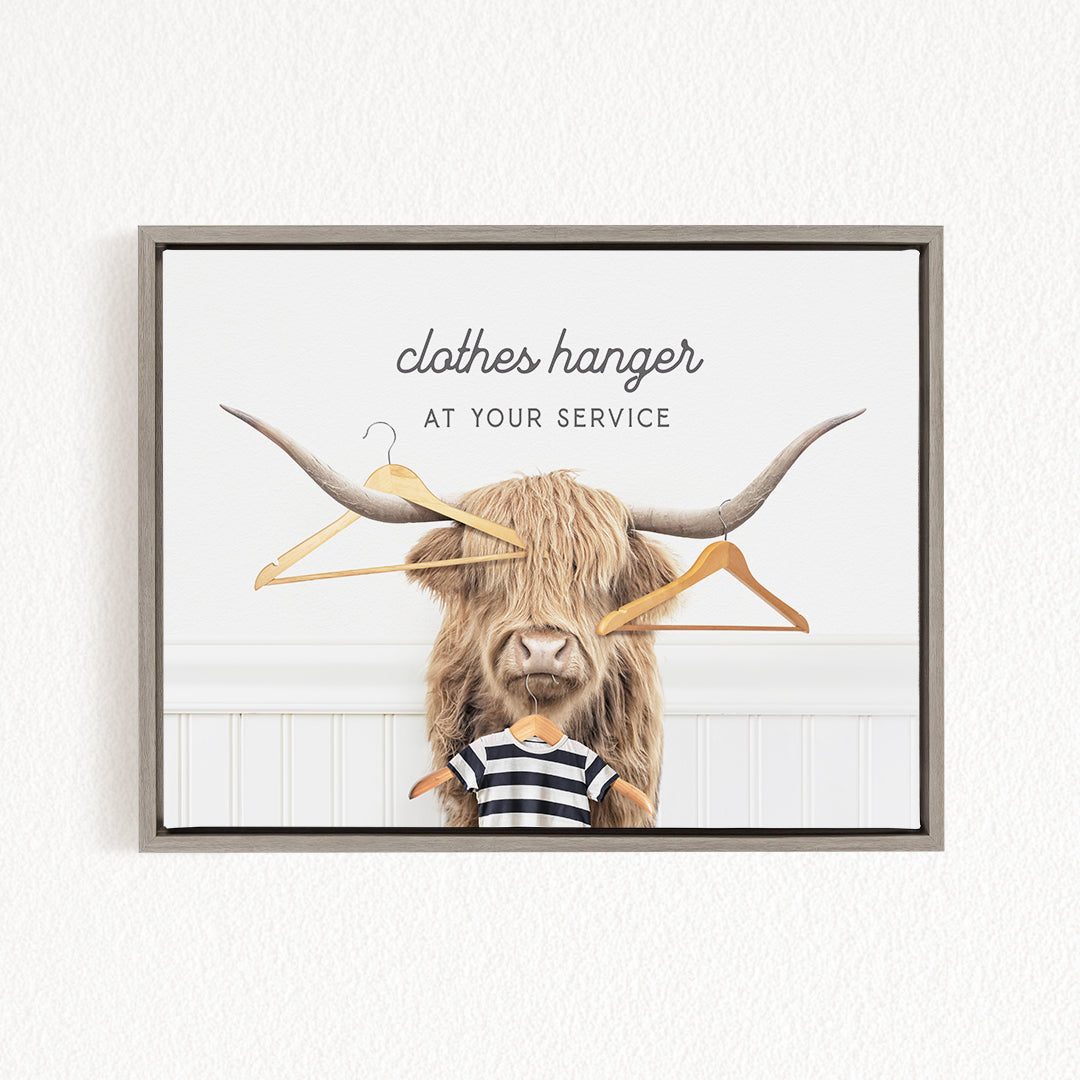 A framed image depicting a horned animal, possibly a yak or a yak dog, holding clothes hangers in its mouth, with the text "clothes hanger at your service" above it.