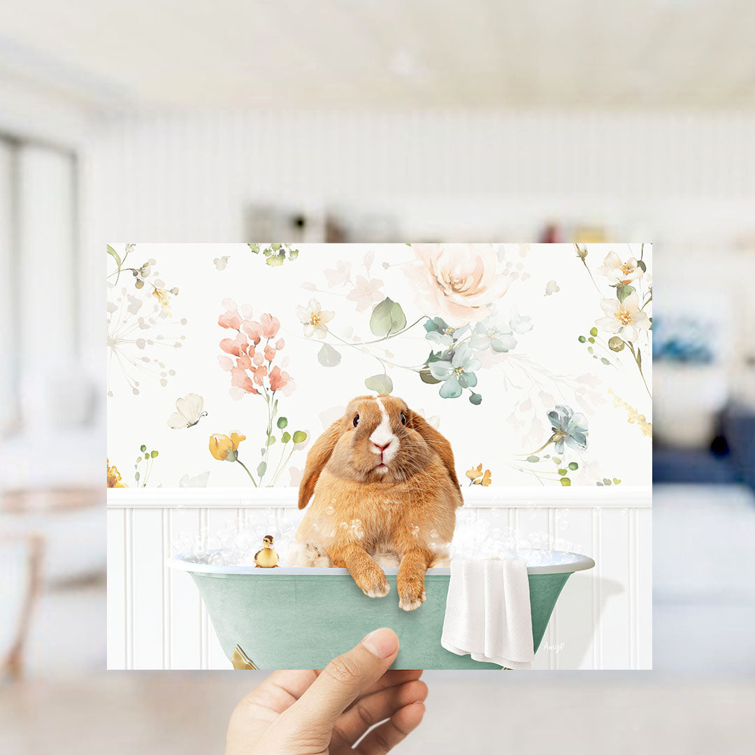 A hand holding a framed image of a brown rabbit sitting in a green bathtub filled with bubbles, surrounded by a floral pattern.
