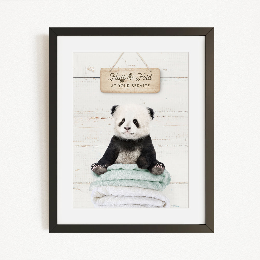 A framed image of a panda bear sitting on a stack of towels, with a wooden sign above that says "Fuffy & Fold at your service".