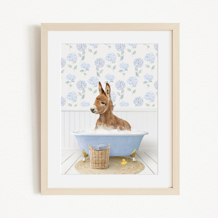 A donkey is sitting in a blue bathtub filled with bubbles, surrounded by a floral patterned wall and a basket of rubber ducks.