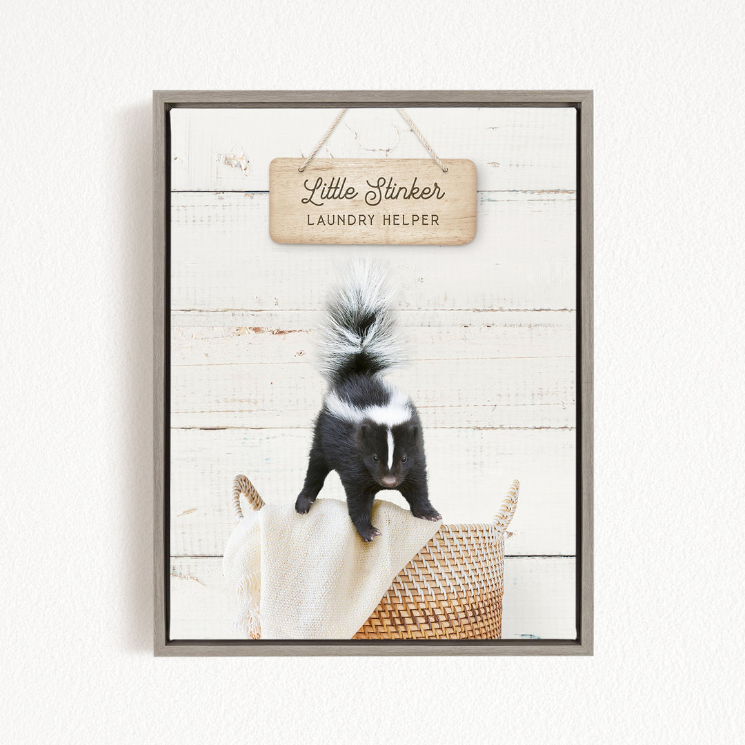 A black and white skunk standing on a woven basket, with a wooden sign above that reads "Little Stinker Laundry Helper".