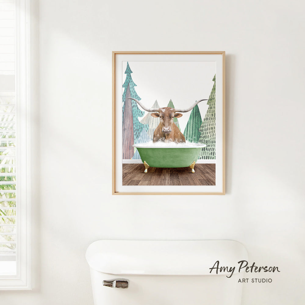 A framed print of a bull with horns sitting in a green bathtub, surrounded by pine trees, is hanging on a white wall.