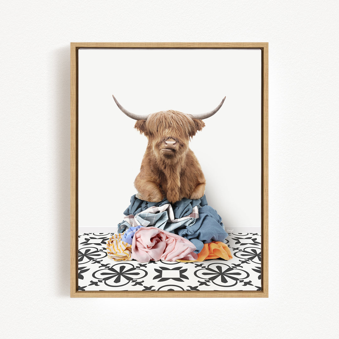 A Highland cow with long horns is sitting atop a pile of clothes, which are scattered on a patterned floor.