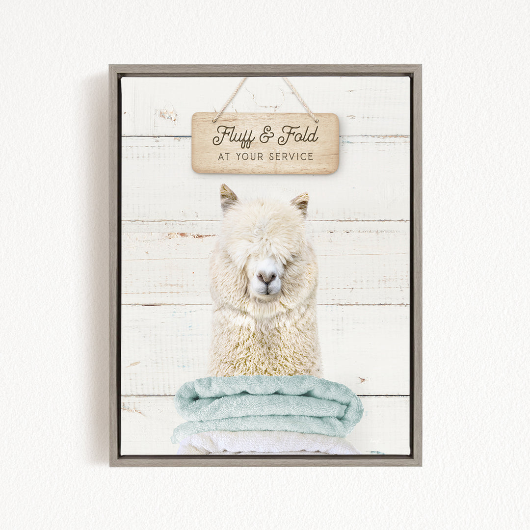 A framed image of a fluffy white alpaca sitting on a stack of blue towels, with a wooden sign above it that says "Fluffy & Fold at your Service".