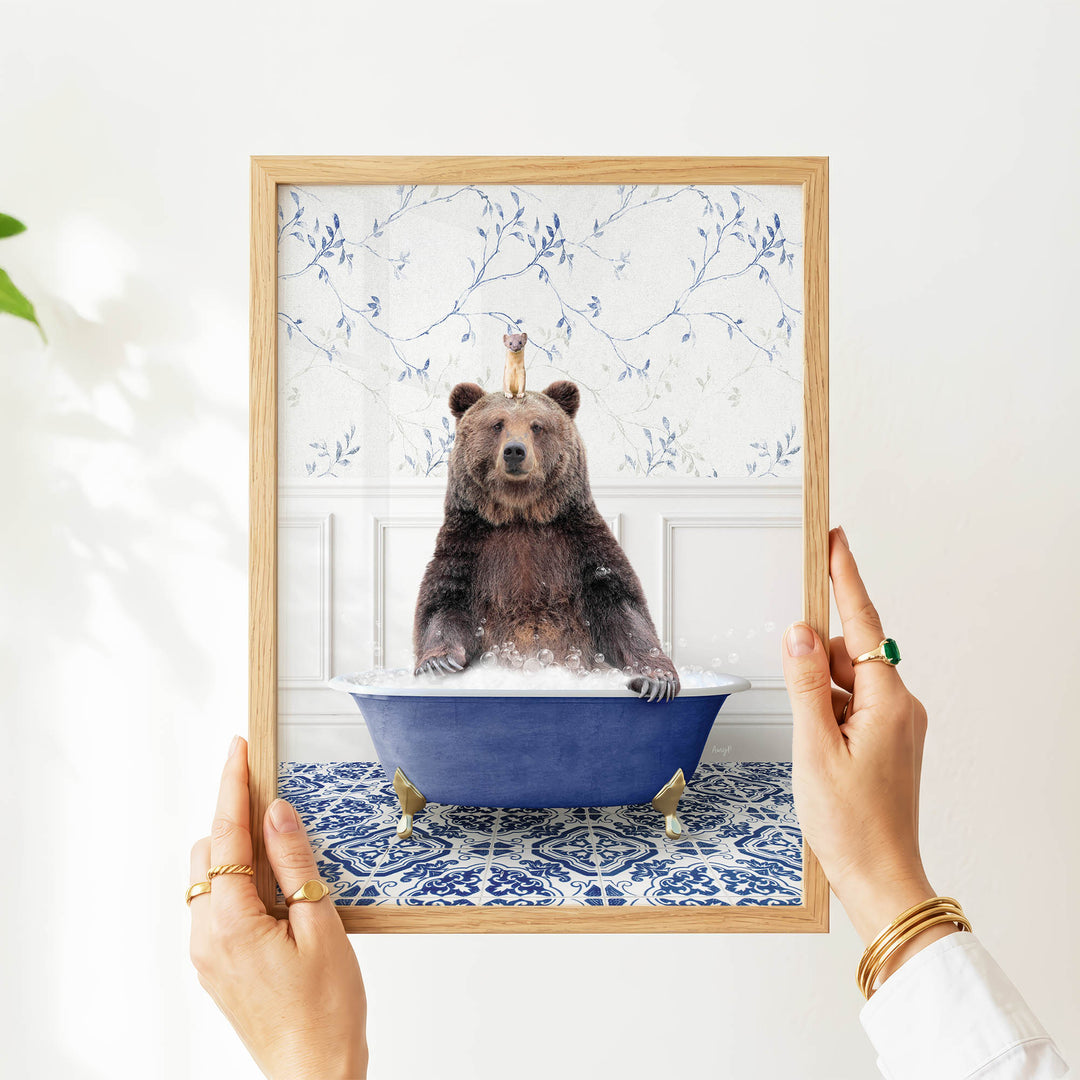 A person's hands are holding a framed picture of a bear sitting in a blue bathtub, with a bird perched on the bear's head.