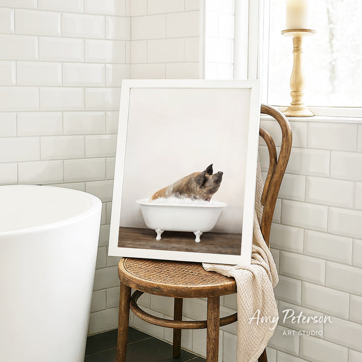 A framed photograph of a dog sitting in a bathtub, with a towel draped over the back of a chair in a bathroom setting.