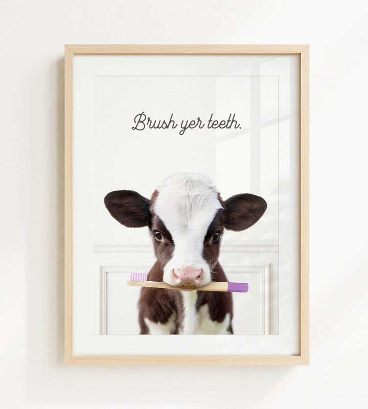 A framed image of a brown and white cow holding a purple toothbrush in its mouth, with the text "Brush yer teeth" written above it.
