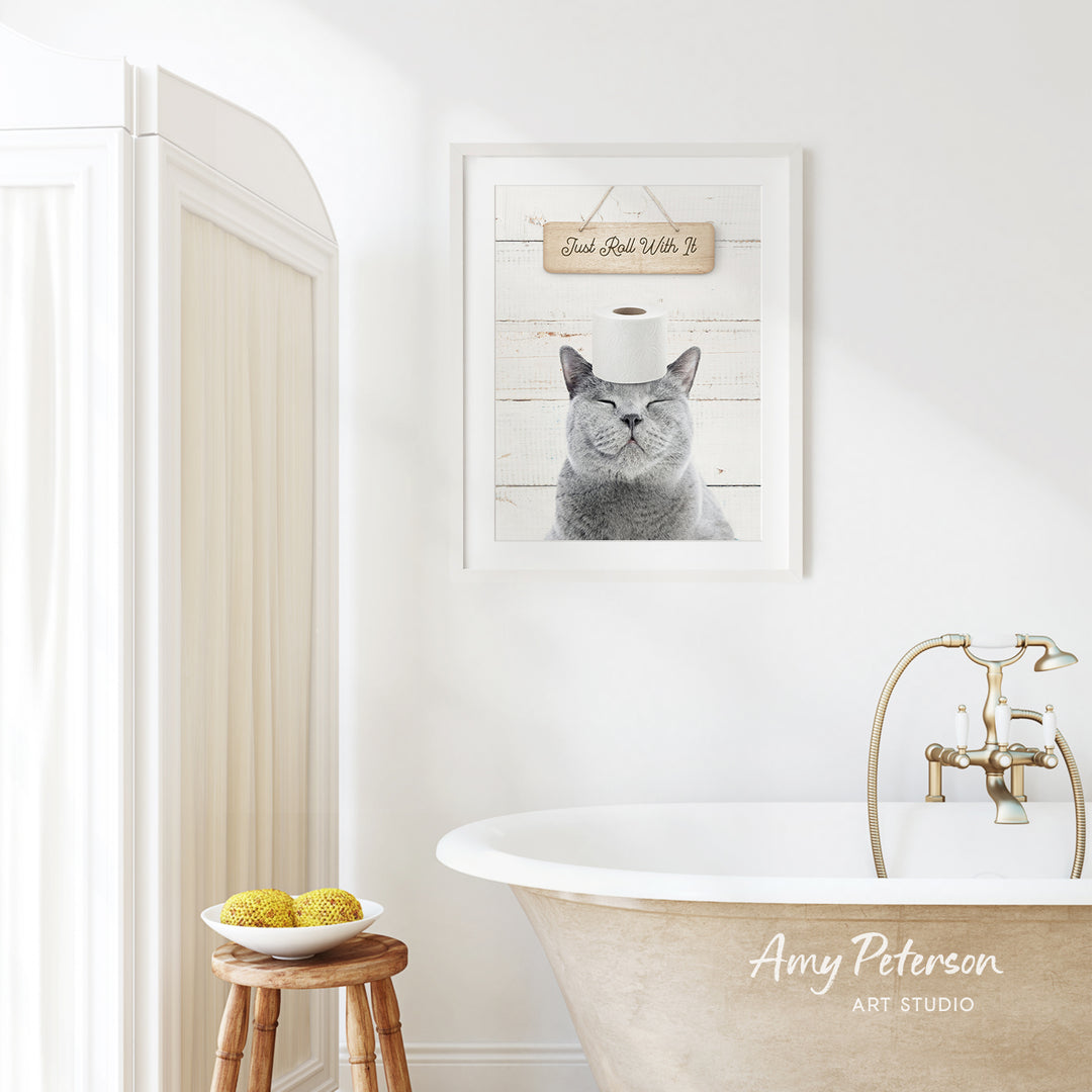 a bathroom with a framed picture of a cat wearing a toilet paper hat, a wooden stool with a bowl of fruit, and a bathtub with a gold faucet.