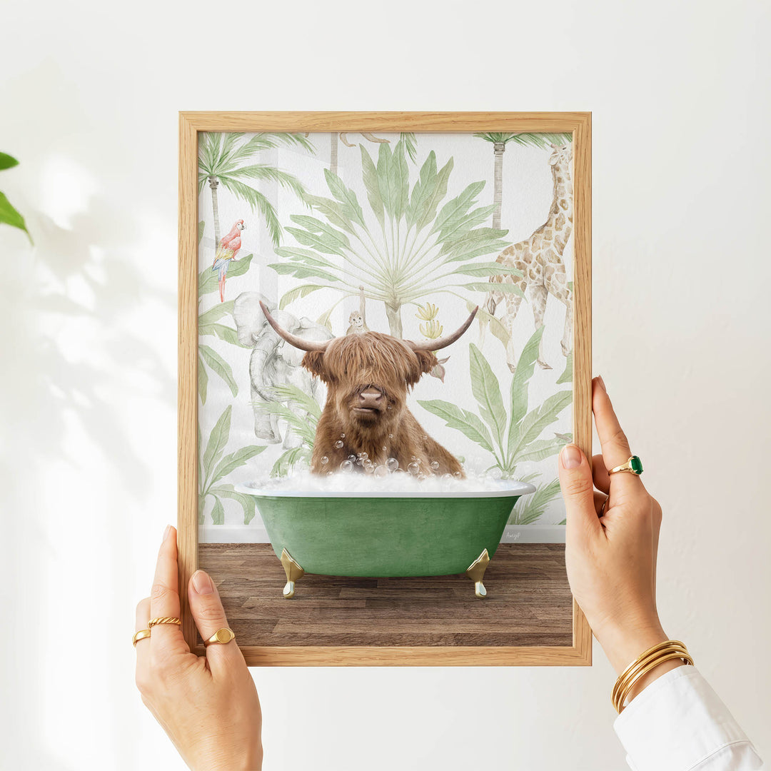 A person's hands are holding a framed picture of a brown cow sitting in a green bathtub, surrounded by tropical foliage.