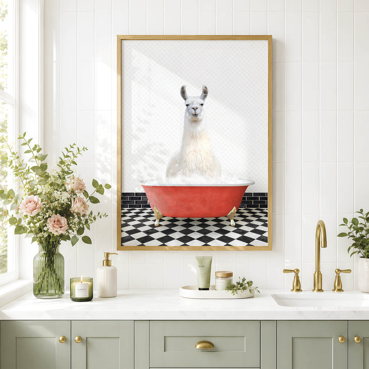 A framed image of a white llama sitting in a red bathtub is displayed on a white tiled wall above a bathroom counter with various toiletries and a vase of flowers.