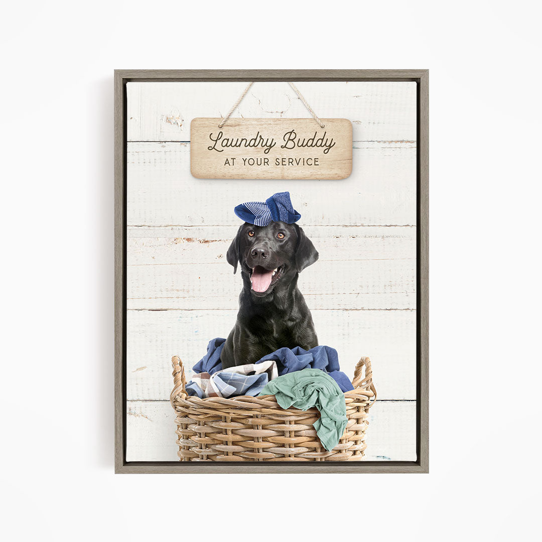 A black dog wearing a blue hat sits in a wicker basket filled with clothes, with a wooden sign above that reads "Laundry Buddy at your service".