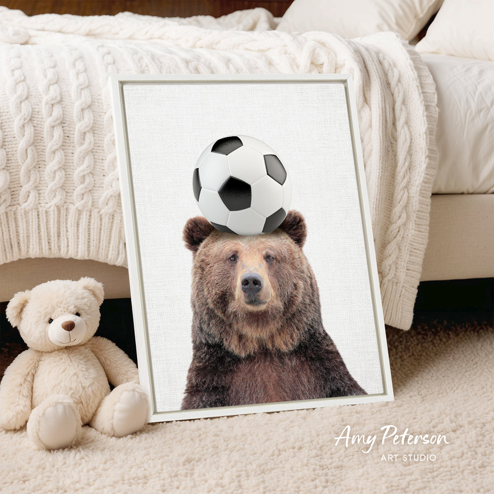 A framed image of a brown bear with a soccer ball on its head, placed on a bed next to a stuffed teddy bear.