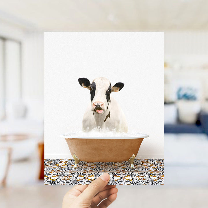 A hand holds up a framed image of a cow sitting in a bathtub filled with water, creating a whimsical and playful scene.