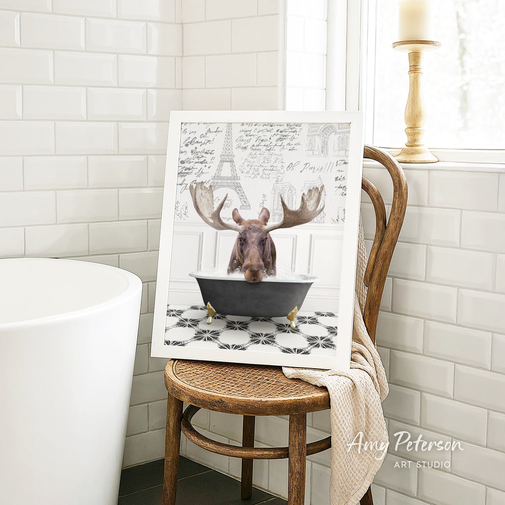 A framed image of a moose sitting in a bathtub, with a towel draped over the back of a wooden chair in a bathroom setting.