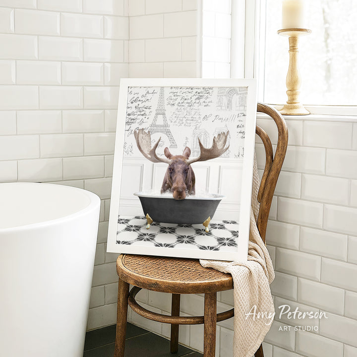 A framed image of a moose sitting in a bathtub, with a towel draped over the back of a wooden chair in a bathroom setting.