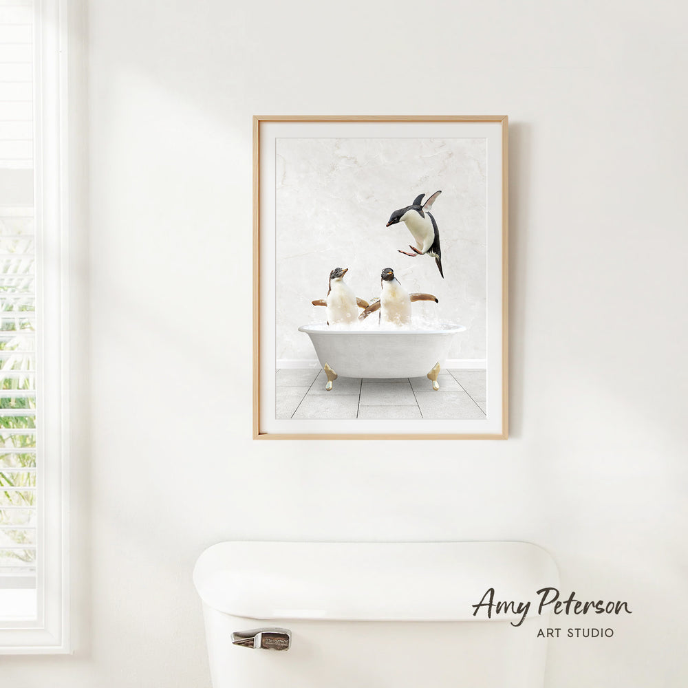 A framed image of two penguins sitting in a bathtub, with one penguin flying towards the other, is hanging on a white wall.