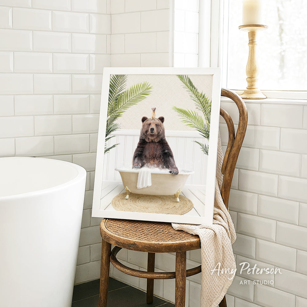 A framed image of a bear sitting in a bathtub, surrounded by palm leaves, is displayed on a wooden chair in a bathroom setting.