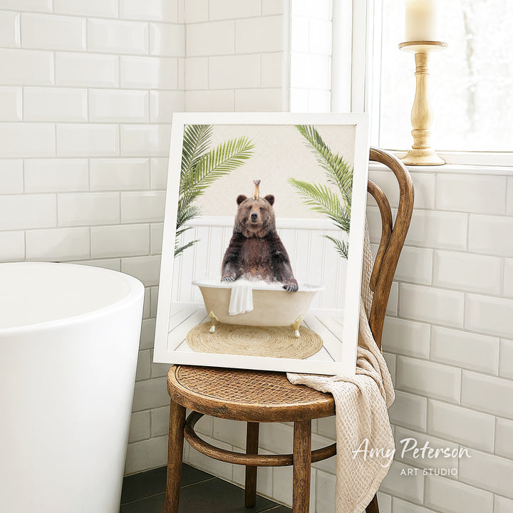 A framed image of a bear sitting in a bathtub, surrounded by palm leaves, is displayed on a wooden chair in a bathroom setting.