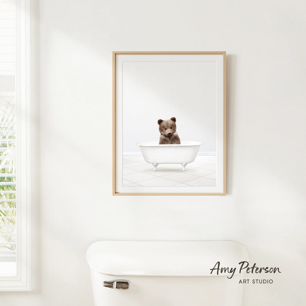 A framed image of a bear sitting in a bathtub is hanging on a white wall.