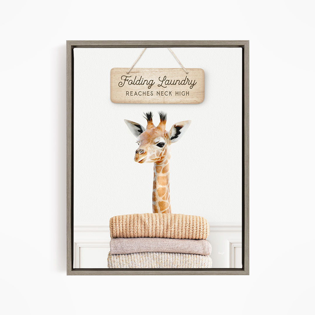 A framed image of a giraffe standing on a pile of folded clothes, with a wooden sign above that says "Folding Laundry Reaches Neck High".