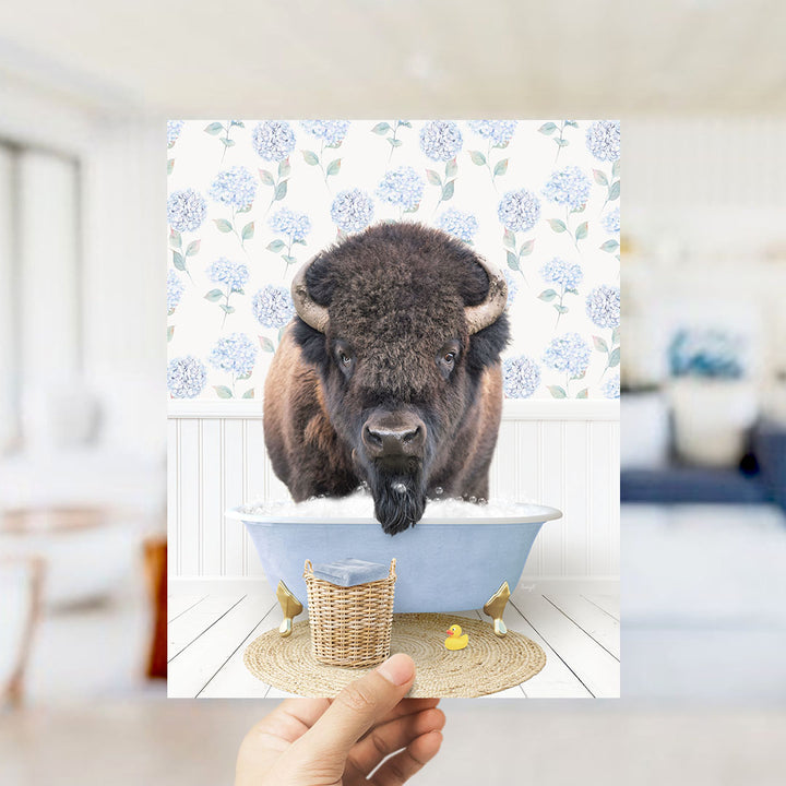 A buffalo is sitting in a bathtub filled with water, surrounded by a blue and white floral patterned wall, a yellow rubber duck, and a woven basket.