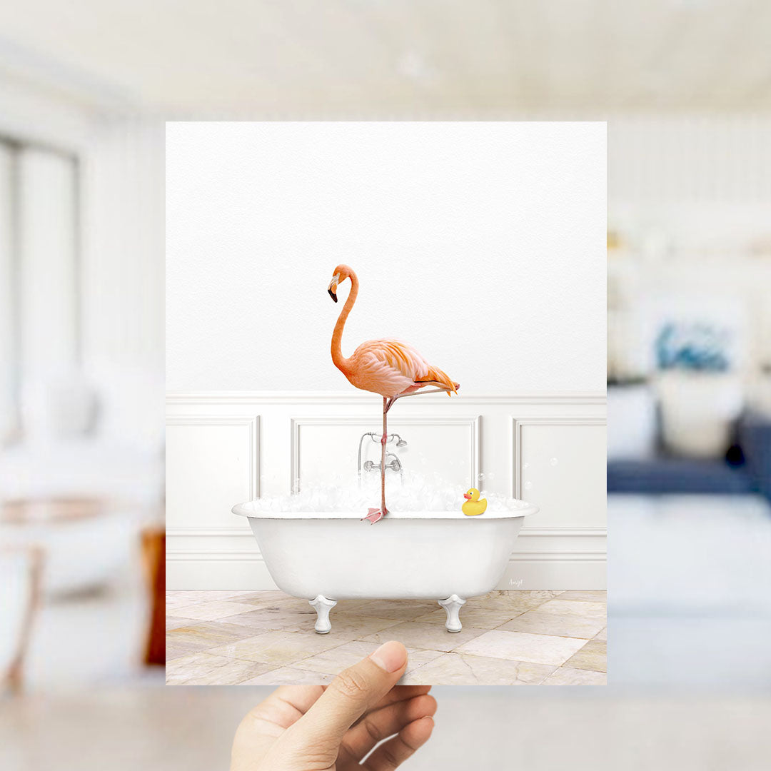 A hand holds up a framed image of a pink flamingo standing on a white bathtub filled with bubbles, with a yellow rubber duck floating nearby.
