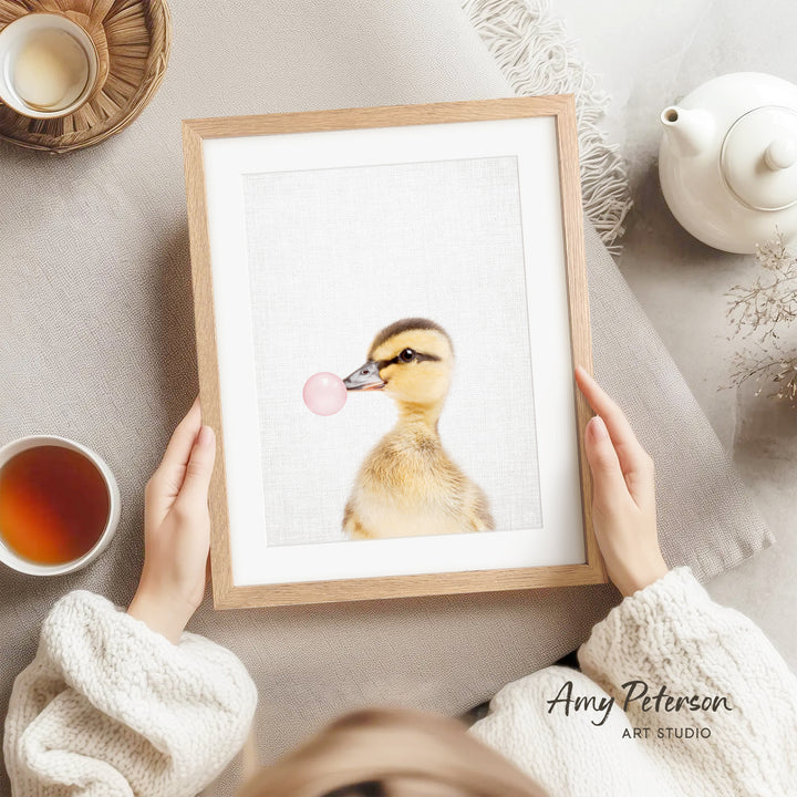 A person is holding a framed picture of a duck blowing a bubble with a pink bubblegum bubble.