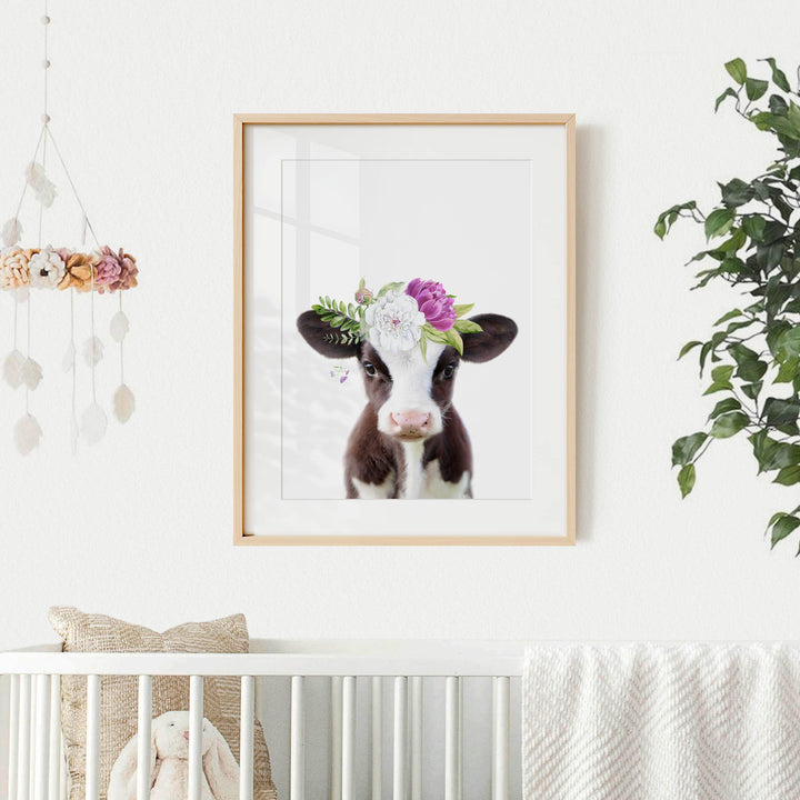 A framed picture of a cow with a floral headpiece hangs on a white wall, accompanied by a hanging mobile and a plant.