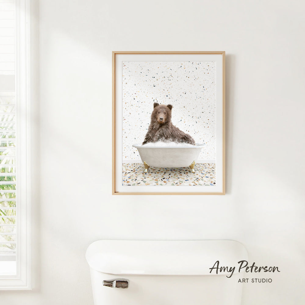 A framed image of a brown bear sitting in a bathtub, surrounded by confetti, is hanging on a white wall.