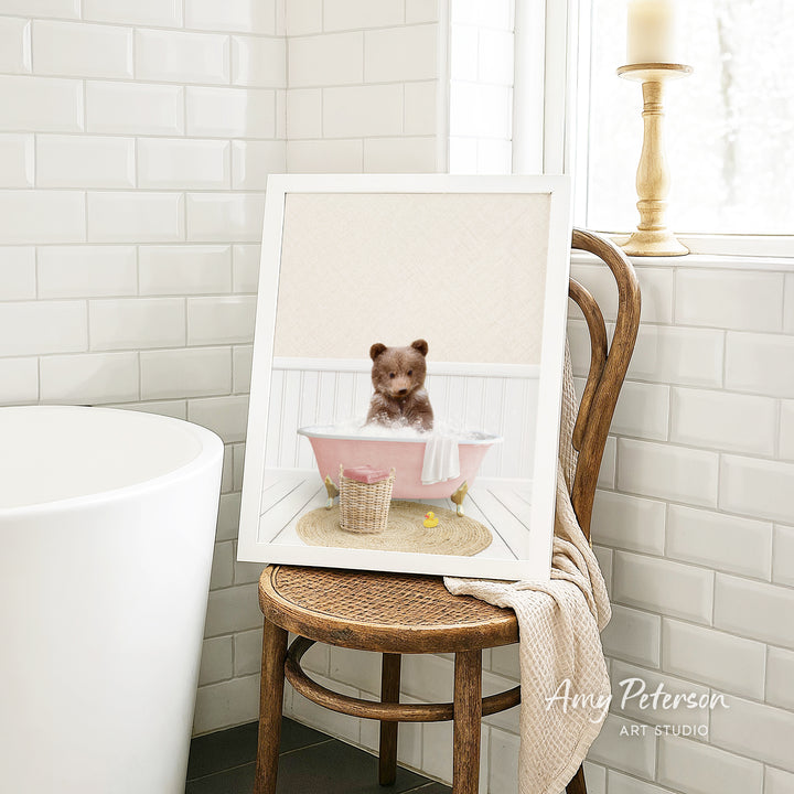 A framed image of a teddy bear sitting in a pink bathtub, surrounded by a basket of bath toys, is displayed on a wooden chair in a bathroom setting.