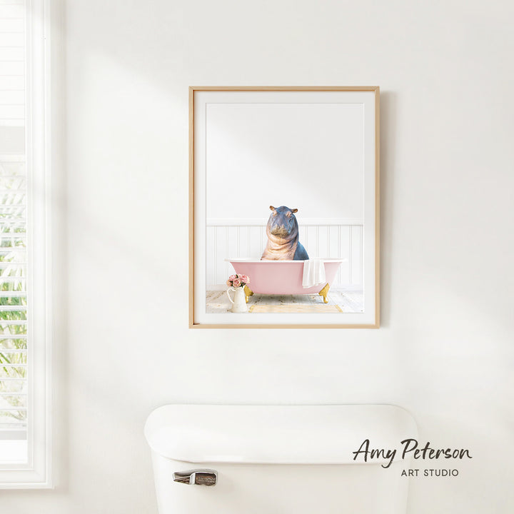 A framed image of a hippo sitting in a pink bathtub, with a window and toilet visible in the background.