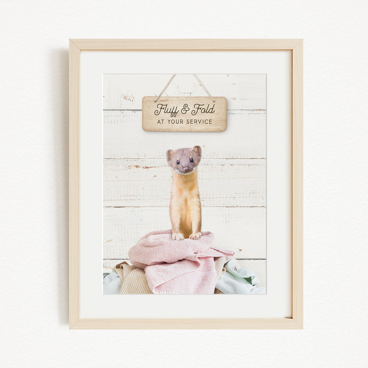 A framed image of a small animal, likely a cat, sitting on a pile of clothes and towels, with a wooden sign above it that says "Fluffy & Fold at your service".