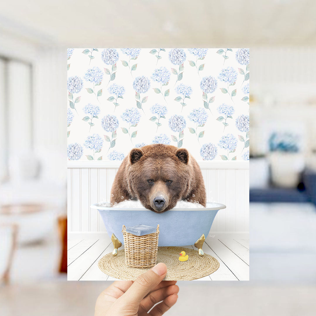 A hand holds up a framed image depicting a brown bear sitting in a blue bathtub filled with bubbles, surrounded by a floral patterned wall.