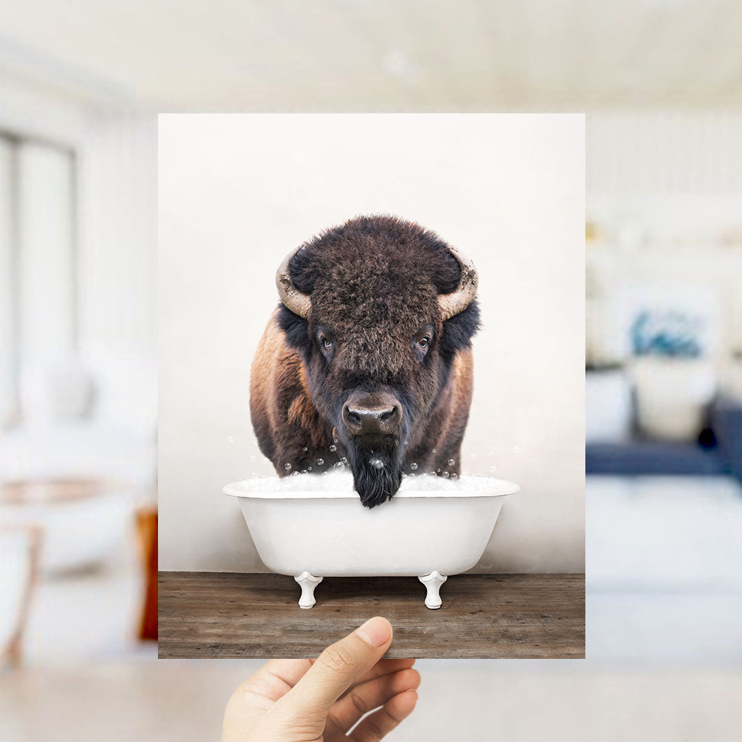 A hand holds up a framed image of a bison with a long beard, standing in a white bathtub.