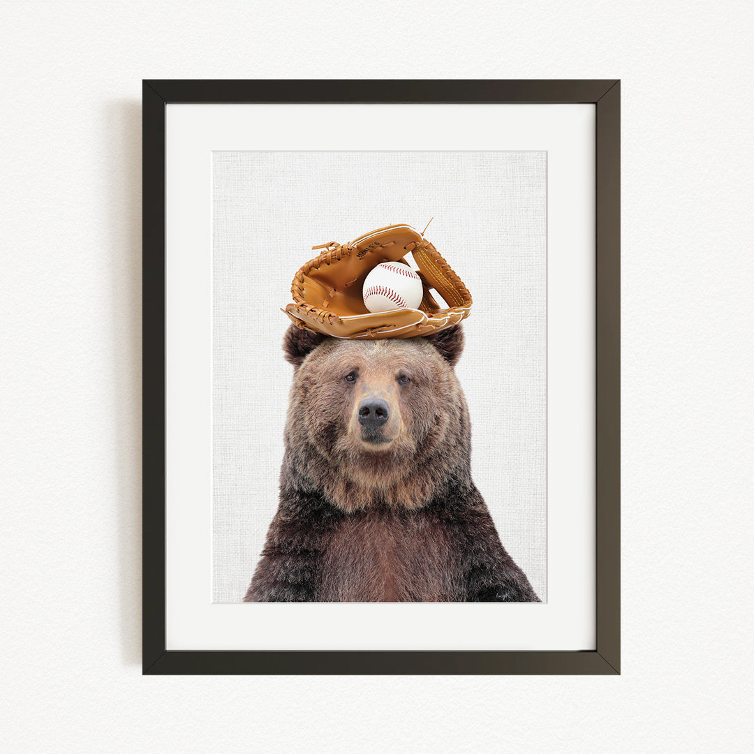 A brown bear wearing a baseball glove on its head, with a baseball resting on top of the glove.