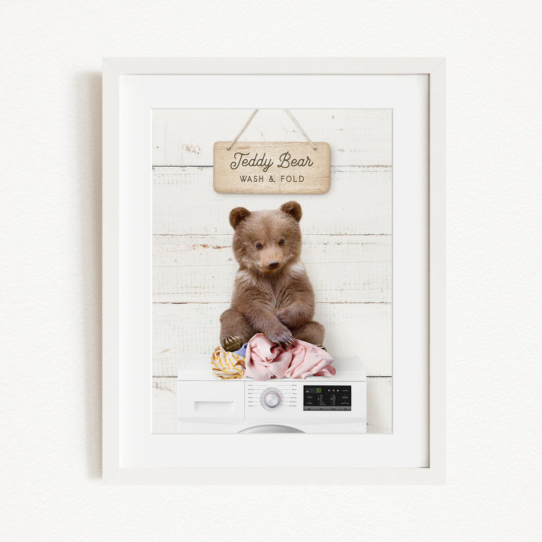 A framed image of a teddy bear sitting on a washing machine, with a sign above it that says "Teddy Bear Wash & Fold".