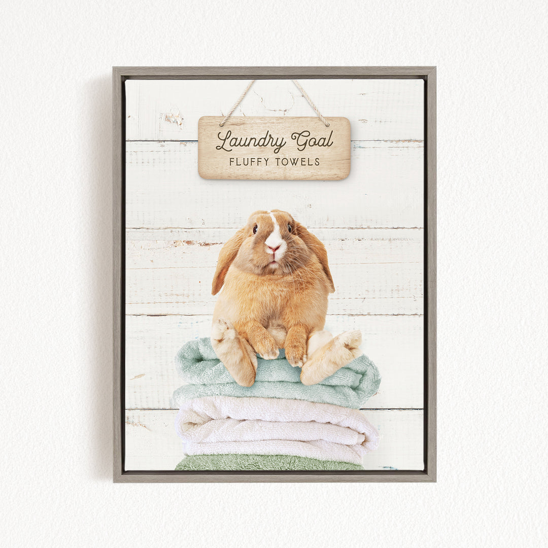 A framed image of a rabbit sitting on a stack of fluffy towels, with a sign above that says "Laundry Goal Fluffy Towels".