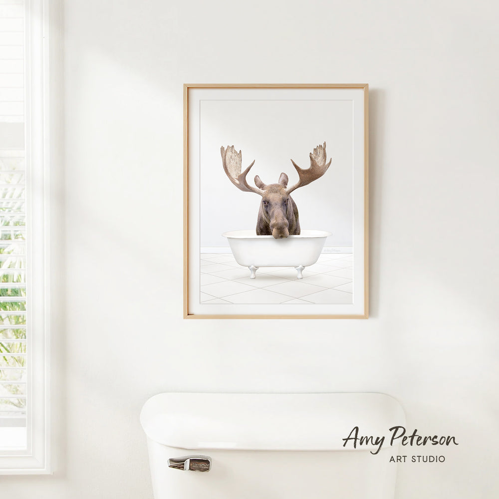 A framed image of a moose sitting in a bathtub is hanging on a white wall.