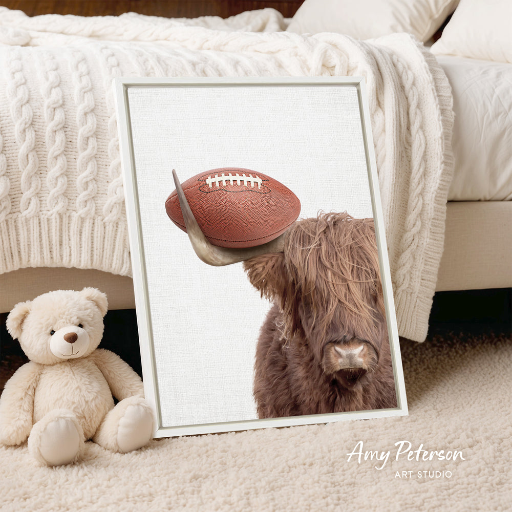 A framed image of a brown cow with a football on its head, placed on a bed next to a teddy bear.