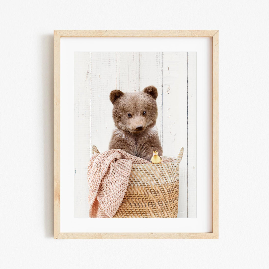 A brown bear sitting in a woven basket, with a pink blanket and a yellow rubber duck nearby.